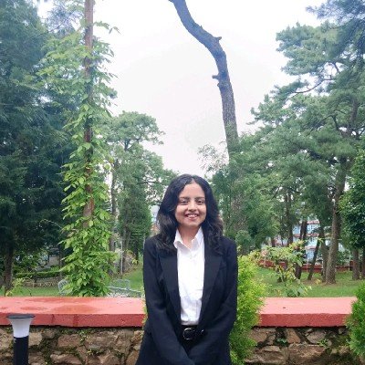 A smiling young woman wearing a black blazer and white shirt sitting in front of a green garden with tall trees in the background